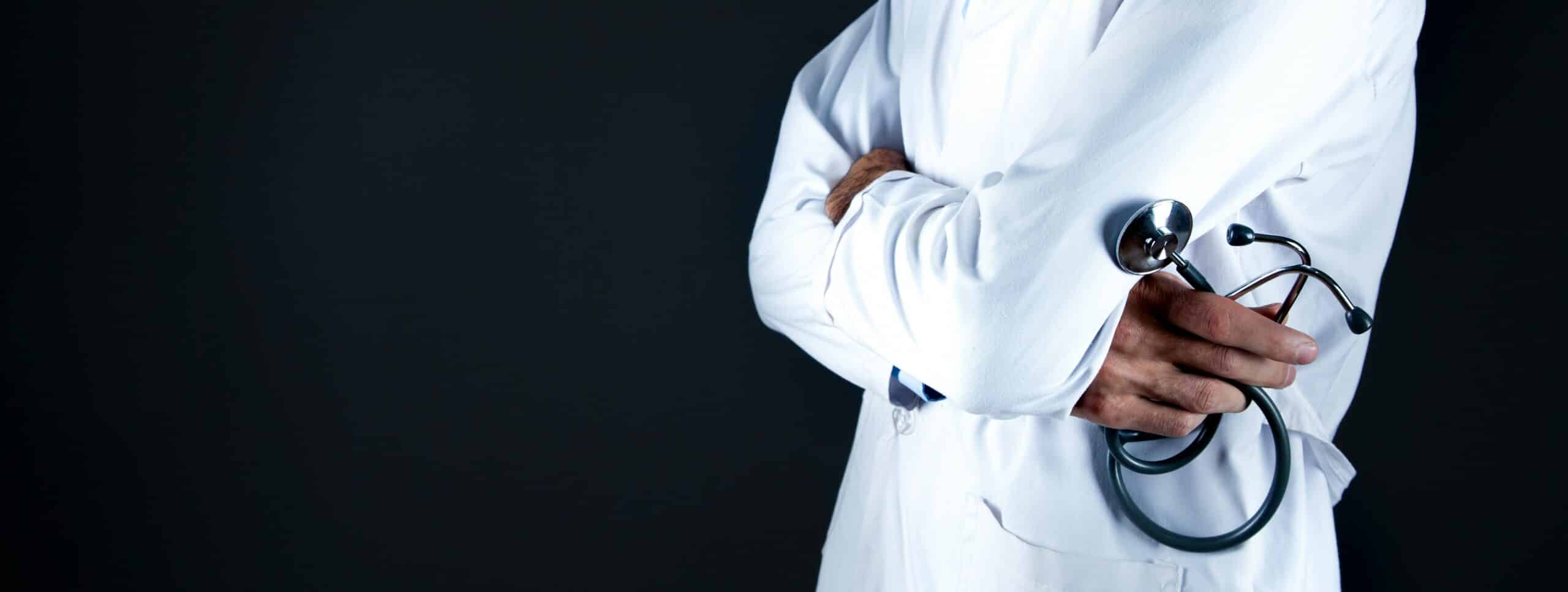 A healthcare professional in a white lab coat with arms crossed, holding a stethoscope, stands against a dark background.