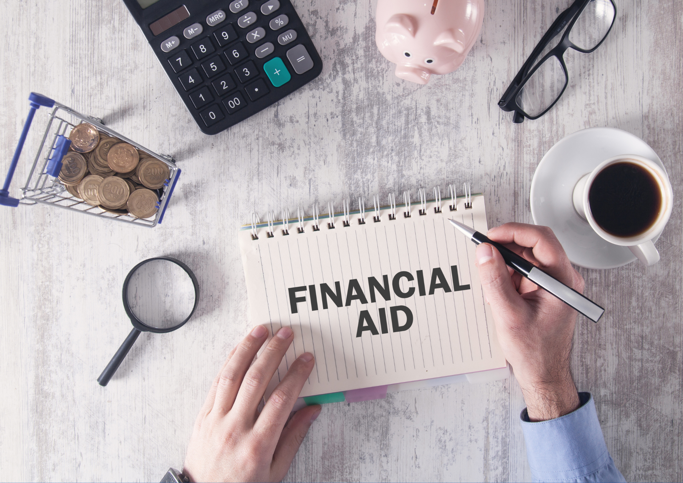 A top-down view of a wooden desk with various items related to financial planning. In the center, an open notebook displays the words "FINANCIAL AID" written in bold black letters, with a hand holding a pen resting on it. Surrounding the notebook are a calculator, a small shopping cart filled with coins, a magnifying glass, a pair of glasses, a pink piggy bank, and a cup of black coffee on a saucer. The scene suggests a focus on budgeting, financial assistance, or financial management.