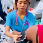 The image shows a medical professional, likely a student or nurse, using a blood pressure cuff to check a patient's blood pressure. The setting appears to be a clinical or community health event, possibly organized by a medical school like All Saints University (ASU), given the context of your previous question. Other individuals in medical attire are visible in the background, suggesting a group effort, perhaps a health outreach program.