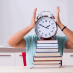 A person wearing a light blue shirt is sitting at a desk with a stack of colorful books in front of them. The person is holding a large, old-fashioned alarm clock with both hands, covering their face, with the clock showing a time close to 12. The background features a modern room with white cabinets, a shelf with books, and a red chair.