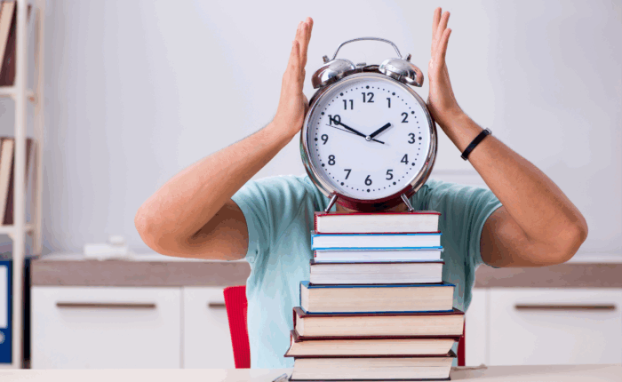 A person wearing a light blue shirt is sitting at a desk with a stack of colorful books in front of them. The person is holding a large, old-fashioned alarm clock with both hands, covering their face, with the clock showing a time close to 12. The background features a modern room with white cabinets, a shelf with books, and a red chair.