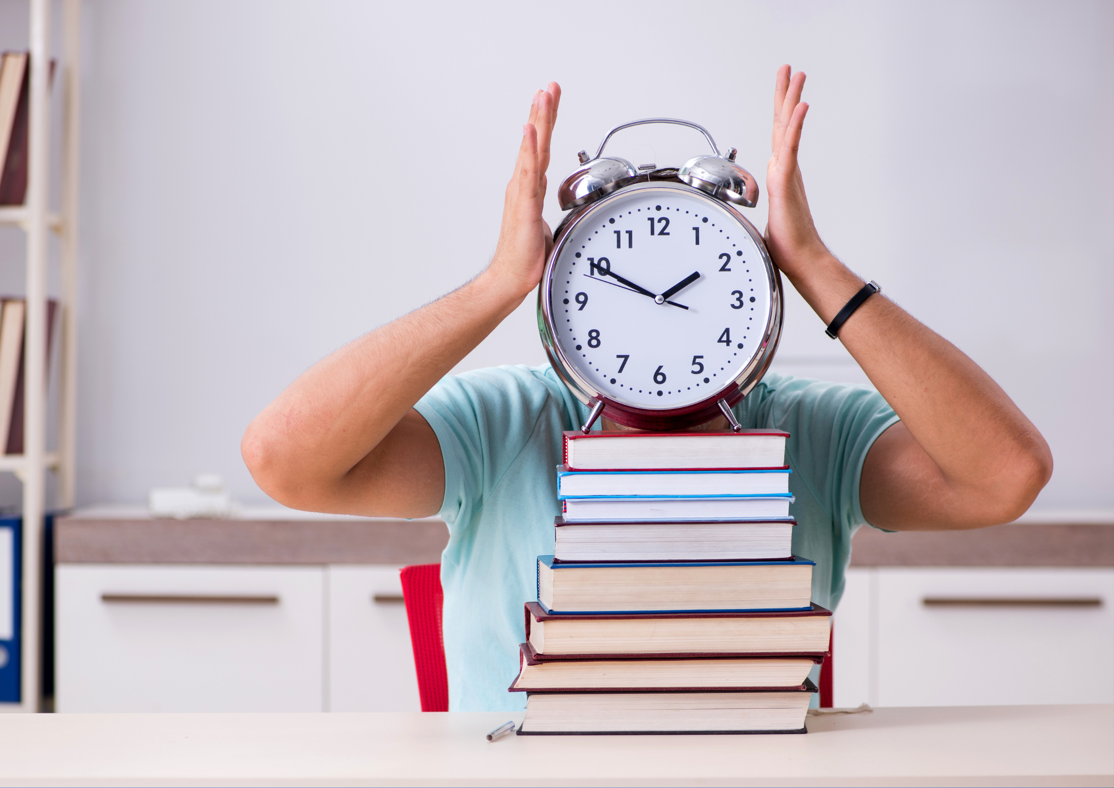 A person wearing a light blue shirt is sitting at a desk with a stack of colorful books in front of them. The person is holding a large, old-fashioned alarm clock with both hands, covering their face, with the clock showing a time close to 12. The background features a modern room with white cabinets, a shelf with books, and a red chair.